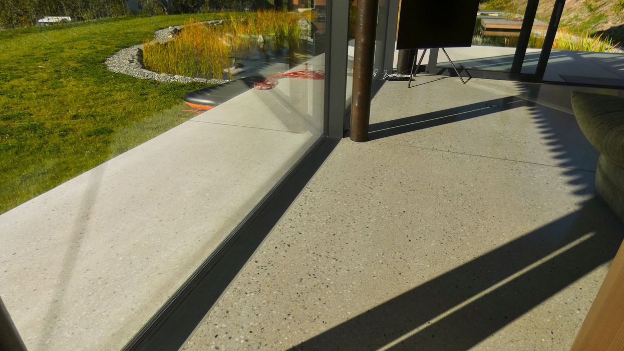 Blick durch eine bodentiefe Glasfront auf eine Terrasse mit hellem Terrazzoboden, davor eine Grünfläche mit einem kleinen Teich.
