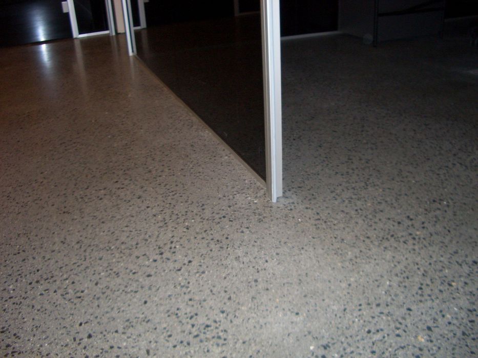 Ein glatter, gesprenkelter Betonboden in einem Innenraum, unterbrochen von einer vertikalen Wandkante in einem dunklen Bereich.