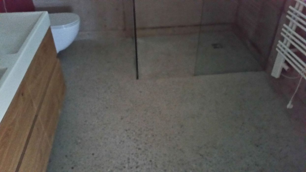 Blick auf ein modernes Badezimmer mit hellem Sichtbetonboden, einem Waschbeckenunterschrank aus Holz, einer weißen Toilette und einer bodengleichen Dusche mit Glaswand.