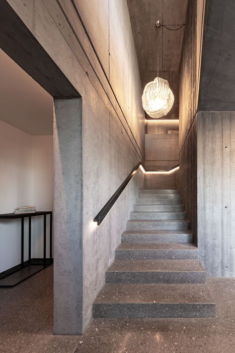 Eine Treppe aus Granit führt in einem modernen Treppenhaus mit Sichtbetonwänden nach oben, beleuchtet von einer dekorativen Hängeleuchte.