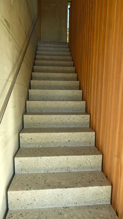 Eine gerade Betontreppe führt zwischen einer Sichtbetonwand mit Handlauf und einer vertikalen Holzlattenwand nach oben.