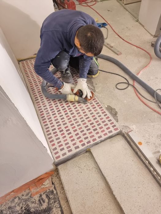 Ein Handwerker kniet auf einer Baustelle und glättet mit einem handgeführten Schleifgerät die Kanten eines verlegten Bodenfliesenmusters.