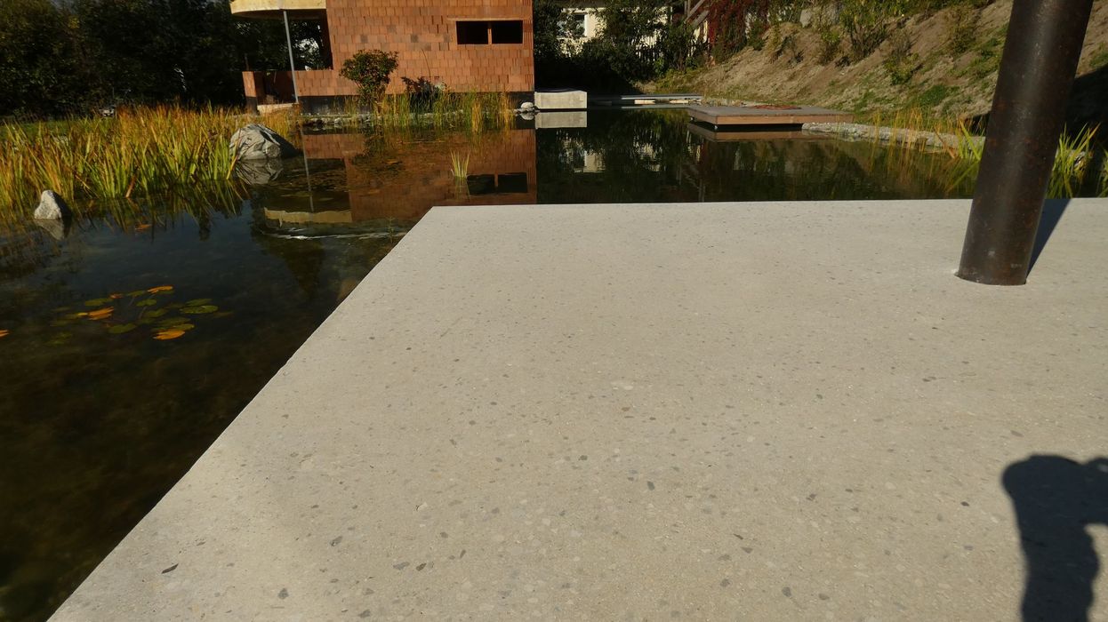 Eine betonierte Terrasse direkt an einem Gartenteich mit einem Rohbau im Hintergrund.