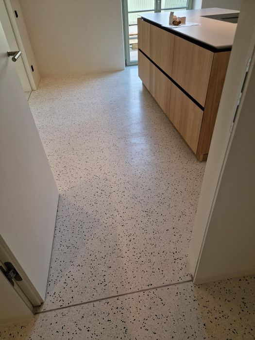 Ein Blick durch eine geöffnete Tür in einen Raum mit hellem Terrazzo-Boden und einem Küchenblock aus hellem Holz.