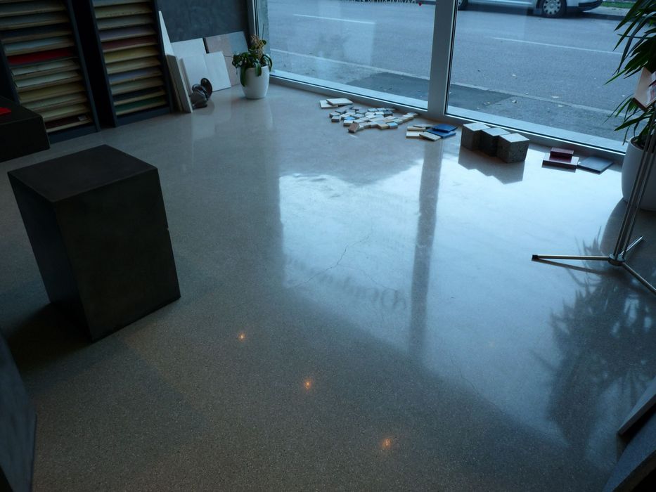 Innenraum eines Showrooms mit einem glänzenden, reflektierenden Betonboden, einem schwarzen Würfel, verschiedenen Bodenfliesenmustern und großen Fenstern mit Blick auf die Straße.