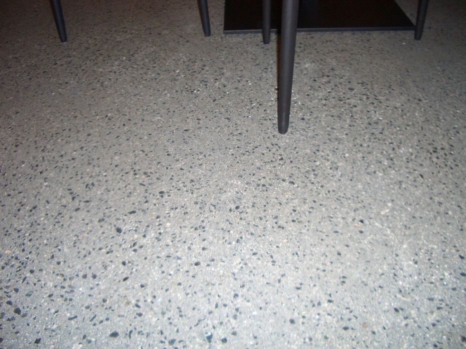 Nahaufnahme eines grauen Terrazzobodens mit kleinen schwarzen und weißen Steinchen, über dem die dunklen Tischbeine eines Möbelstücks zu sehen sind.