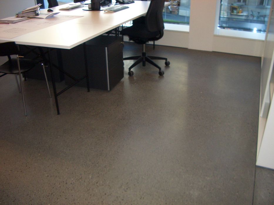 Blick in ein modernes Büro mit einem weißen Schreibtisch, schwarzem Bürostuhl und einem anthrazitfarbenen Betonboden.