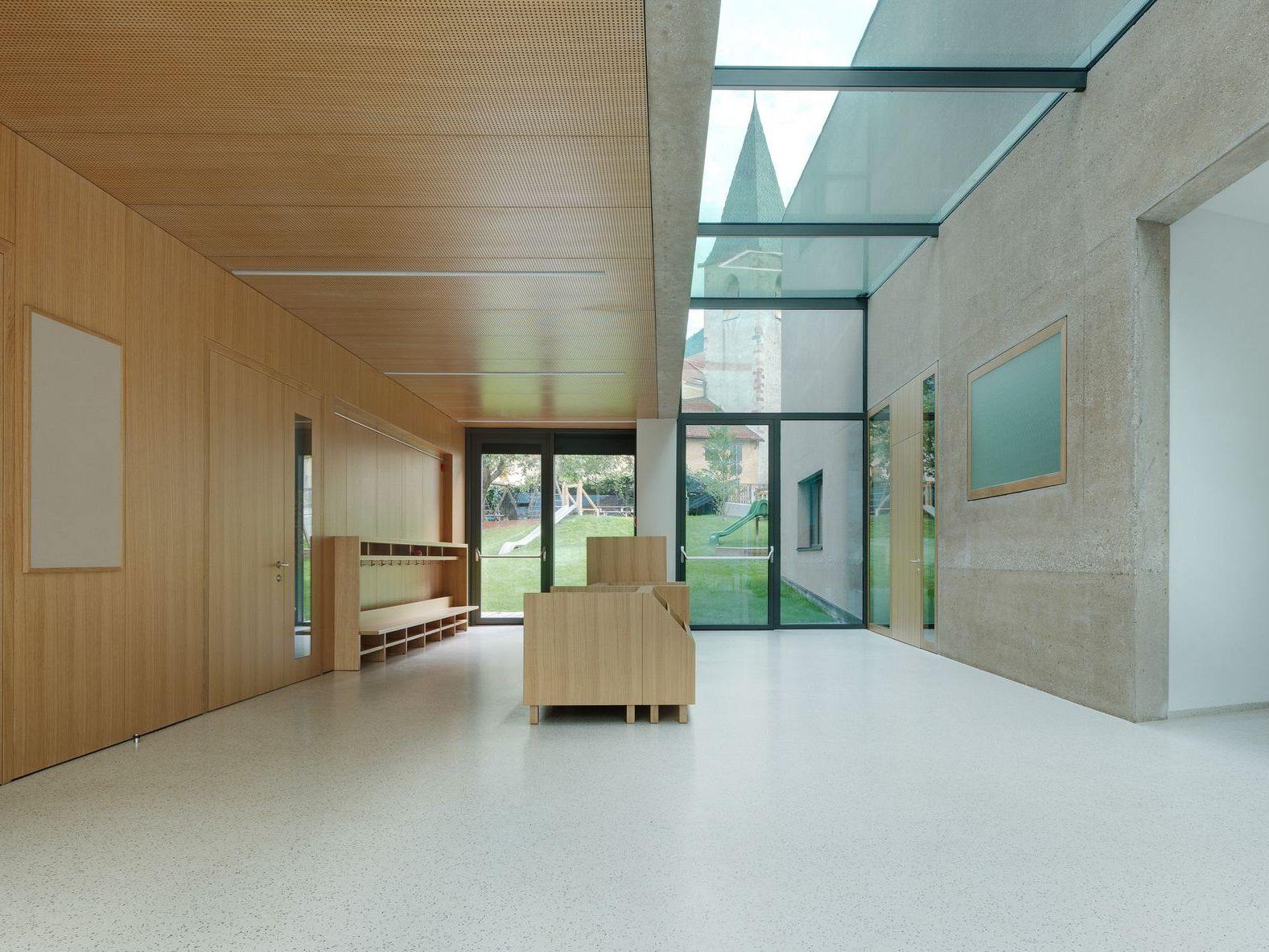 Ein heller Flur in einem modernen Gebäude mit Holzwand, Glasdach und Blick auf einen Spielplatz sowie einen Kirchturm.