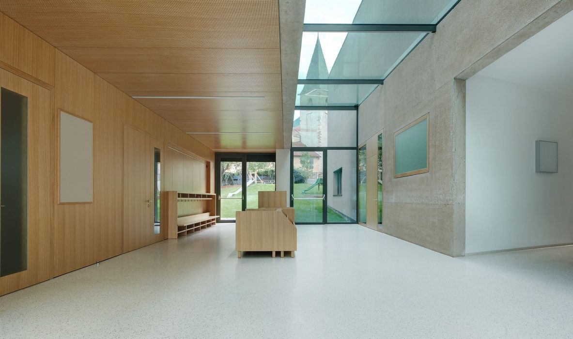 Ein heller Flur in einem modernen Gebäude mit Holzwand, Glasdach und Blick auf einen Spielplatz sowie einen Kirchturm.