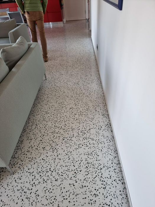 Ein heller Flur mit einem Terrazzoboden, auf dem eine Person im grünen Oberteil und beigefarbener Hose neben einem grauen Sofa steht.