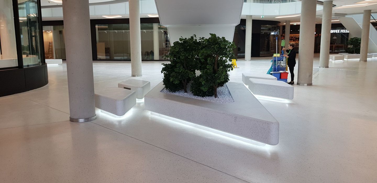 Ein heller, moderner Innenbereich eines Einkaufszentrums mit minimalistischen, beleuchteten Sitzbänken aus Stein, umgeben von kleinen Bäumen in einem Pflanzbeet, während im Hintergrund eine Reinigungskraft arbeitet.