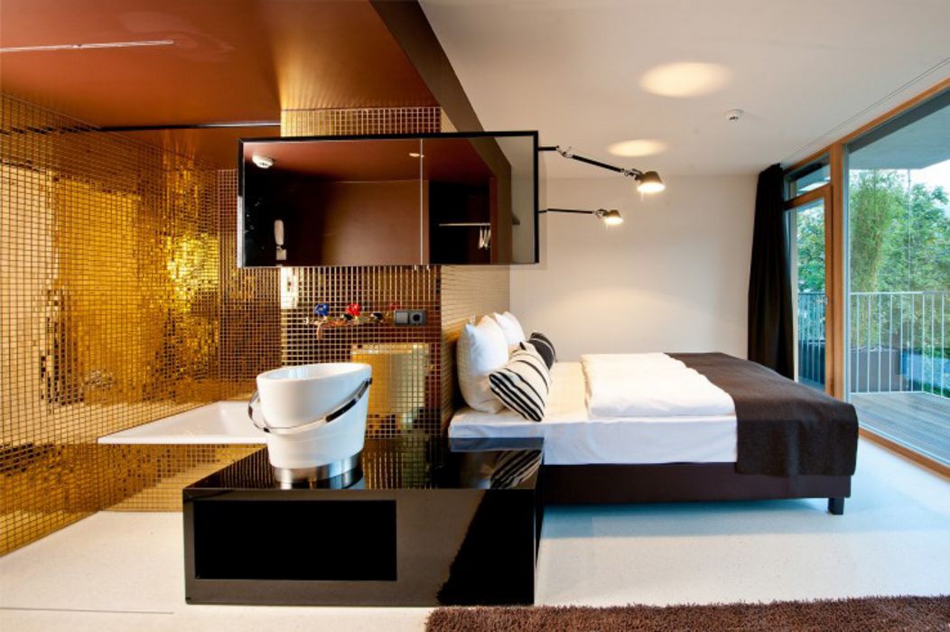Modernes Hotelzimmer mit direkt angrenzendem Badezimmer, das durch goldene Mosaikfliesen besticht, sowie einem Doppelbett neben einem Balkon mit Grünblick.