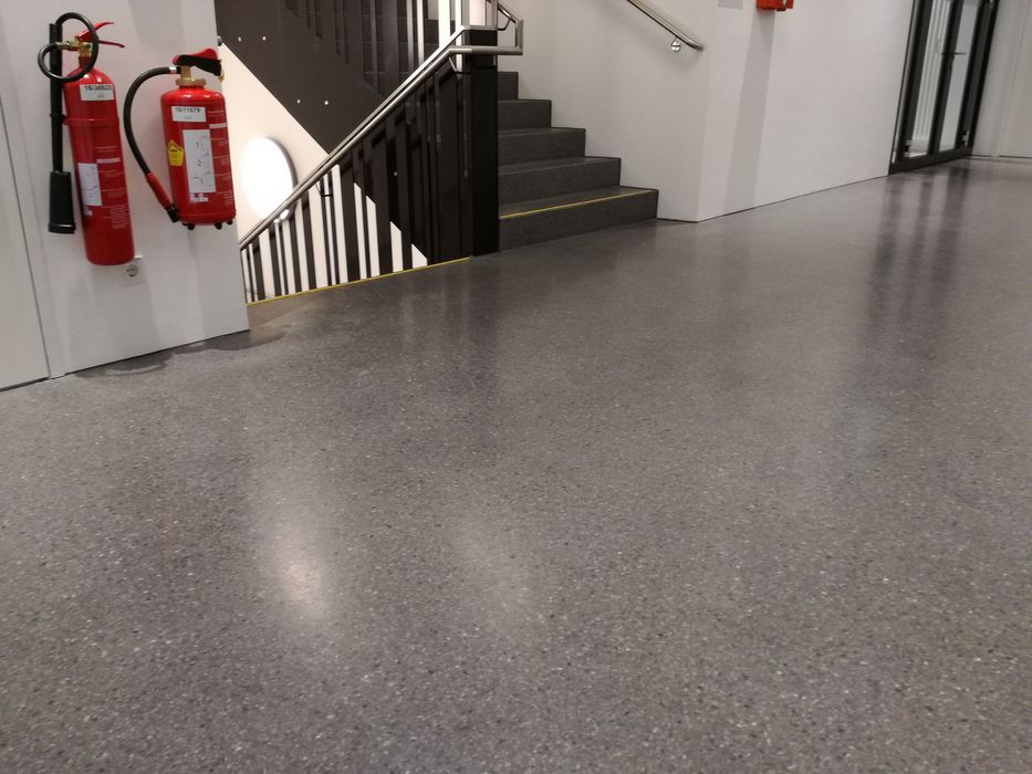 Zwei rote Feuerlöscher hängen an einer weißen Wand neben einem Treppenaufgang mit dunklen Stufen und einem silbernen Geländer in einem modernen Gebäude.