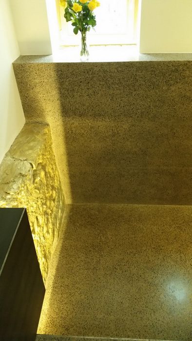 Ein Blick in einen Raum mit einer Wand aus Natursteinmauerwerk, die indirekt beleuchtet wird, und einer Glasvase mit gelben Rosen auf einem Fensterbrett.