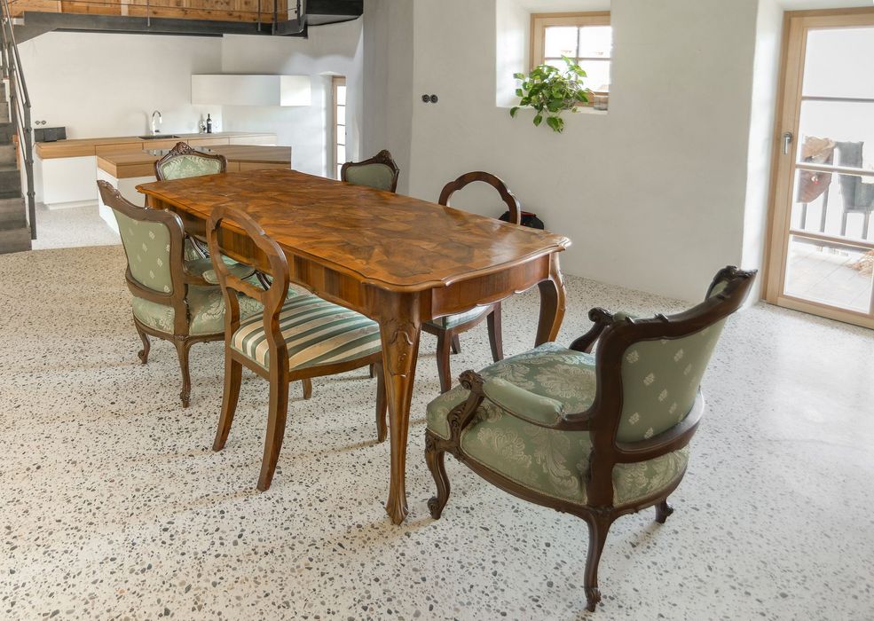 Ein antiker Holztisch mit vier verzierten Stühlen mit grünem Stoffbezug steht in einem Raum mit hellem Terrazzoboden, im Hintergrund ist eine moderne Küche zu sehen.