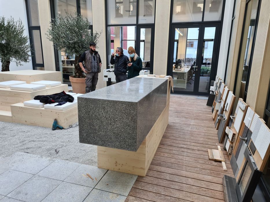 Drei Personen stehen in einem Innenhof mit einem großen Natursteinblock-Tresen, Holzmöbeln, Olivenbäumen und Materialmustern an der Wand.
