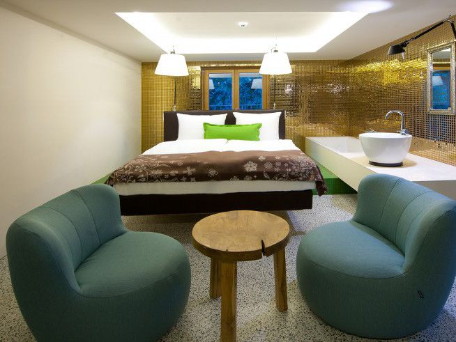 Ein modernes Hotelzimmer mit einem Doppelbett, zwei türkisfarbenen Sesseln, einem kleinen Holztisch und einer goldenen Mosaikwand, an der ein Waschbecken angebracht ist.