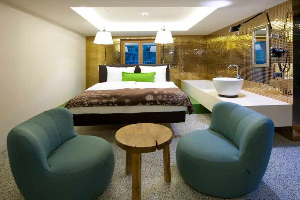 Ein modernes Hotelzimmer mit einem Doppelbett, zwei türkisfarbenen Sesseln, einem kleinen Holztisch und einer goldenen Mosaikwand, an der ein Waschbecken angebracht ist.