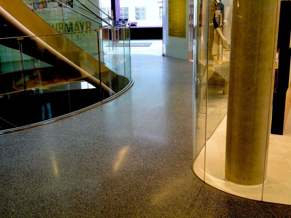 Ein modern gestalteter Innenraum mit einer geschwungenen Glastreppe und einem runden Betonpfeiler, der als Schaufensterfront dient.