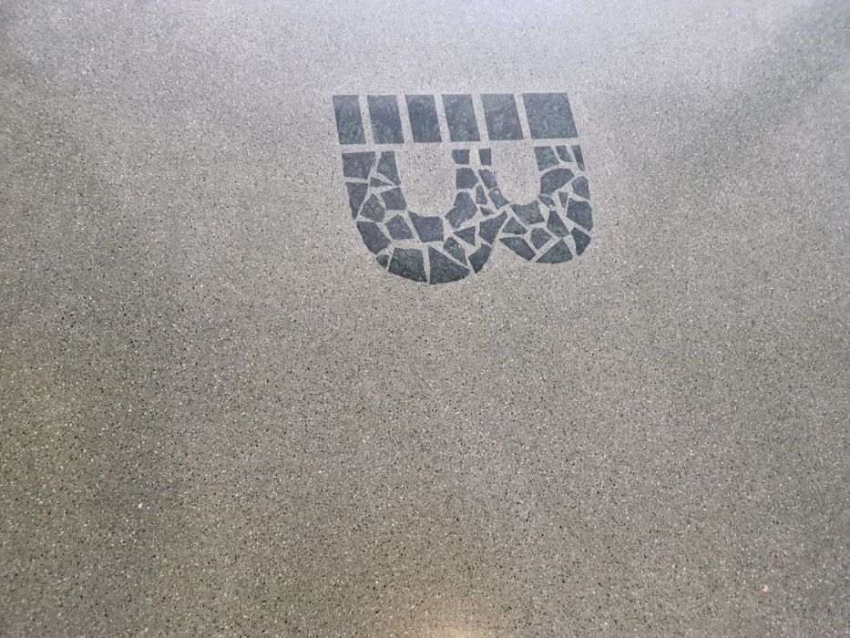 Ein grauer Betonboden mit einem in den Boden eingelassenen Logo in Form eines stilisierten Buchstabens aus dunklen Mosaiksteinen.