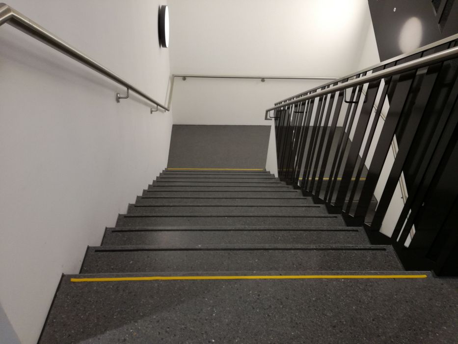 Blick von oben auf eine moderne Treppe mit dunkelgrauen Stufen, die jeweils mit einem gelben Sicherheitsstreifen markiert sind sowie ein Handlauf auf der linken und ein Metallgeländer auf der rechten Seite.