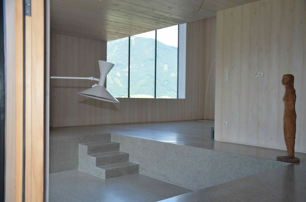 Ein minimalistisch gestalteter Innenraum mit hellen Holzwänden, einer Steinstufe und einem großen Fenster mit Bergblick, ergänzt durch eine moderne Wandleuchte und eine kleine Holzskulptur.