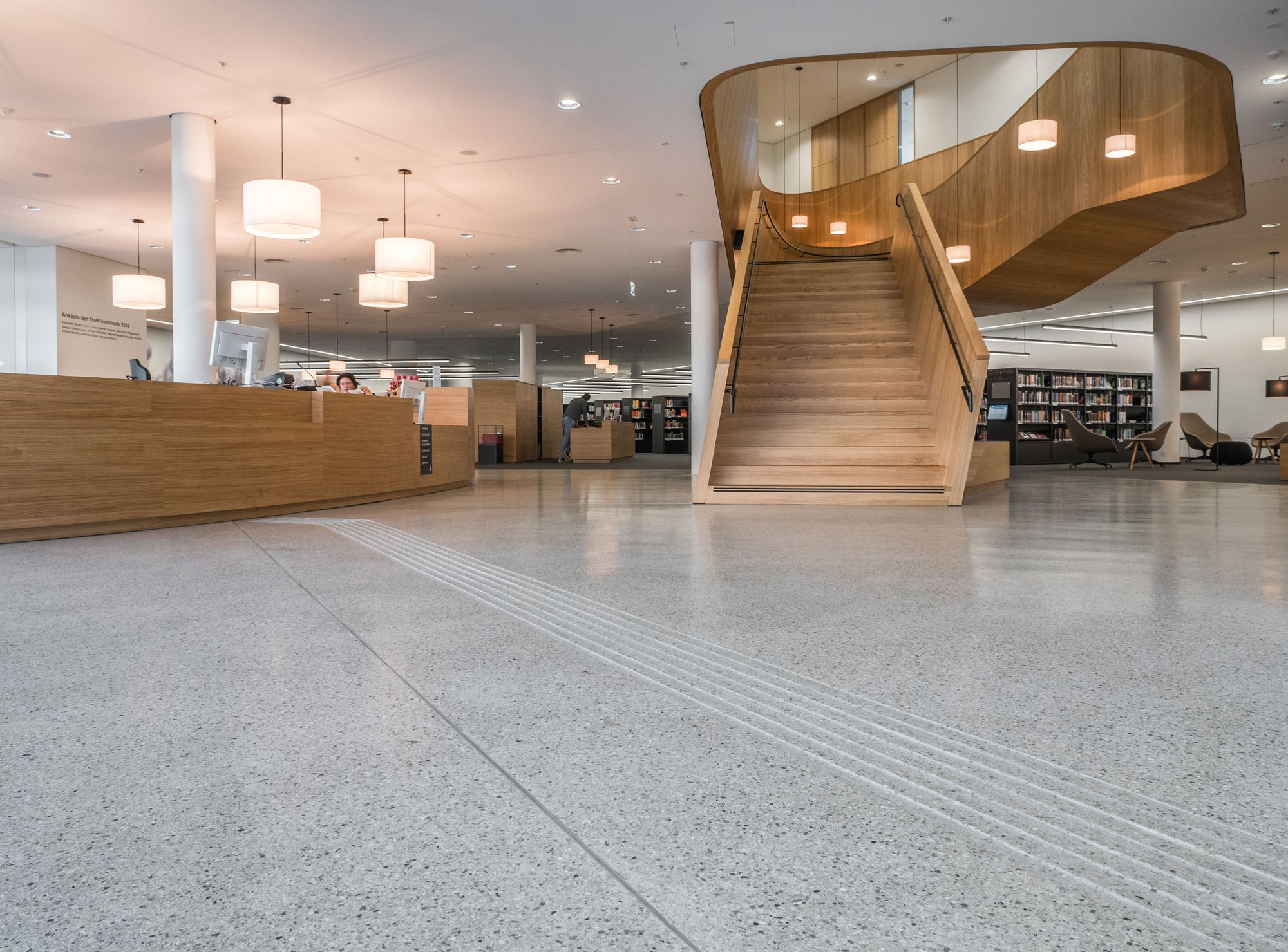 Innenansicht einer modernen Bibliothek mit einem hellen Terrazzoboden, einer geschwungenen Holztreppe, einem langen Empfangstresen aus Holz und mehreren weißen Pendelleuchten.